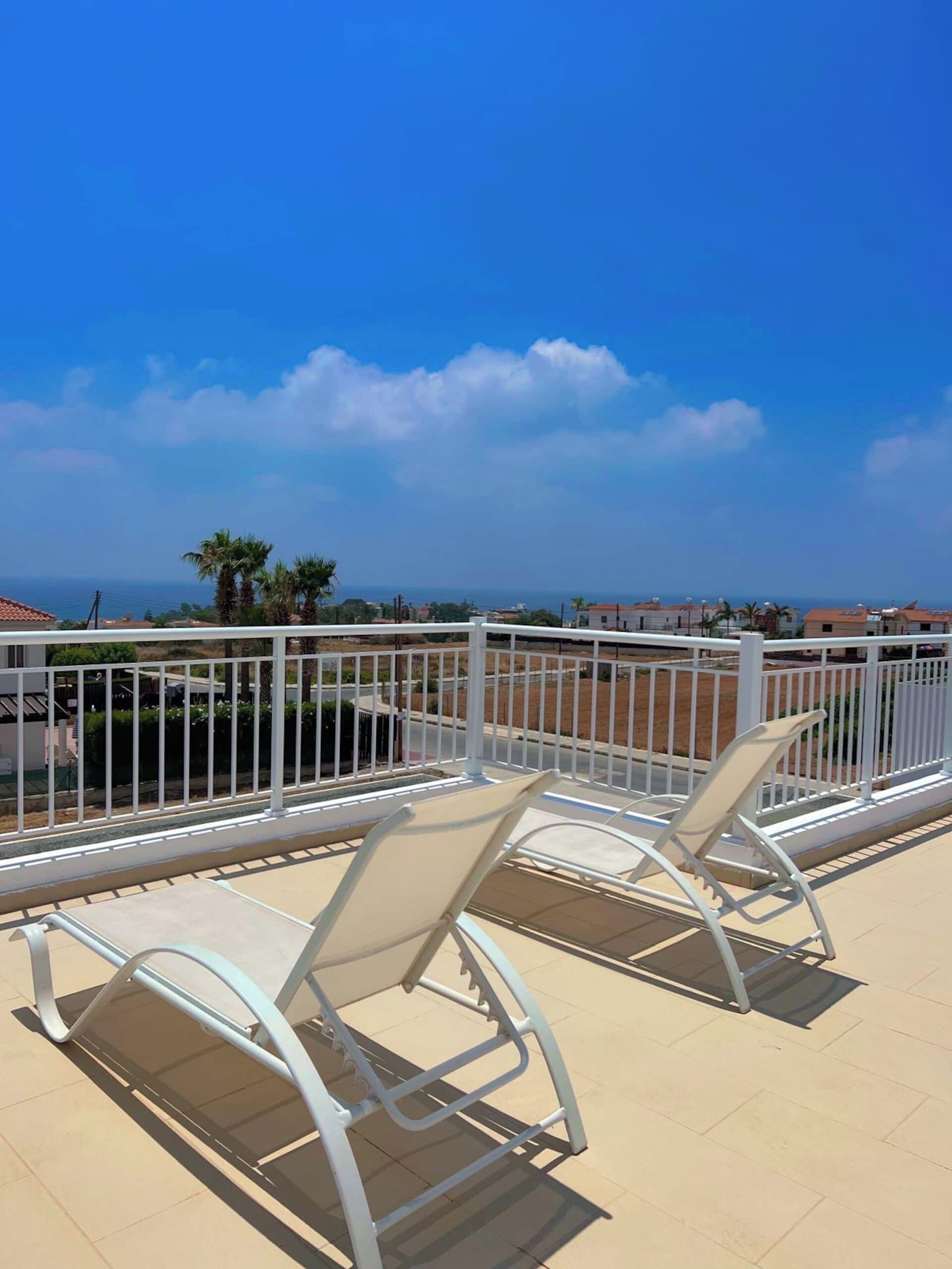 A balcony with two white lounge chairs, a white railing, and a view of the sea and buildings in the distance.
