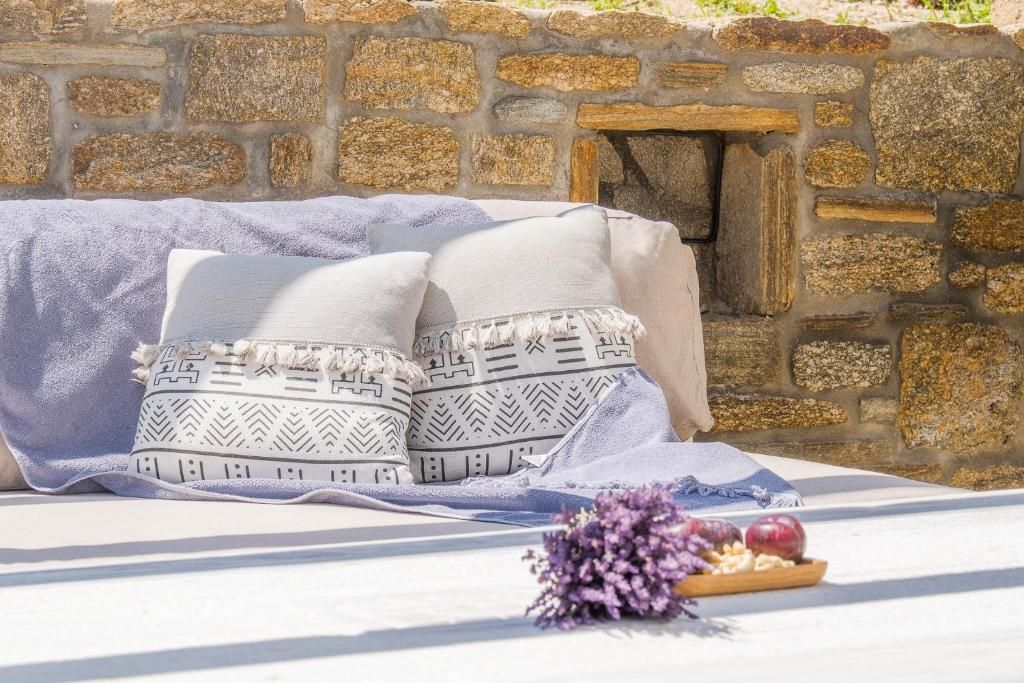 A stone wall, a bed with a white sheet and two pillows, and a small table with a bouquet of flowers and a bowl of fruit.