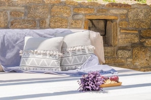 A stone wall, a bed with a white sheet and two pillows, and a small table with a bouquet of flowers and a bowl of fruit. - Ventus Villa (MYKONOS)