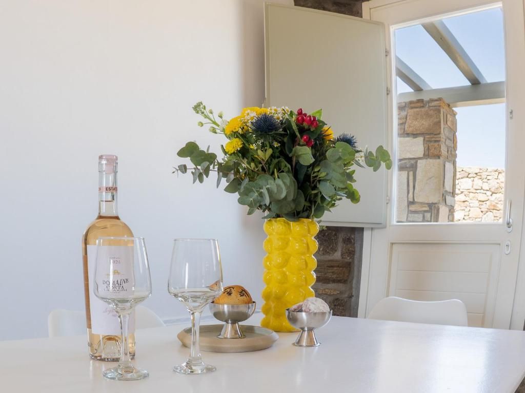 A table with a bottle of wine, two glasses, a bowl of bread, and a yellow vase with flowers.