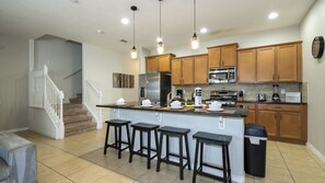 A modern kitchen with wooden cabinets, a central island with stools, stainless steel appliances, and pendant lighting. - 4BR Townhome Solterra Resort Lazy River & Pool (Davenport)