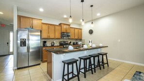 A modern kitchen with a stainless steel refrigerator, a microwave, and a coffee maker. There are wooden cabinets and a large island with bar stools. - 4BR Townhome Solterra Resort Lazy River & Pool (Davenport)