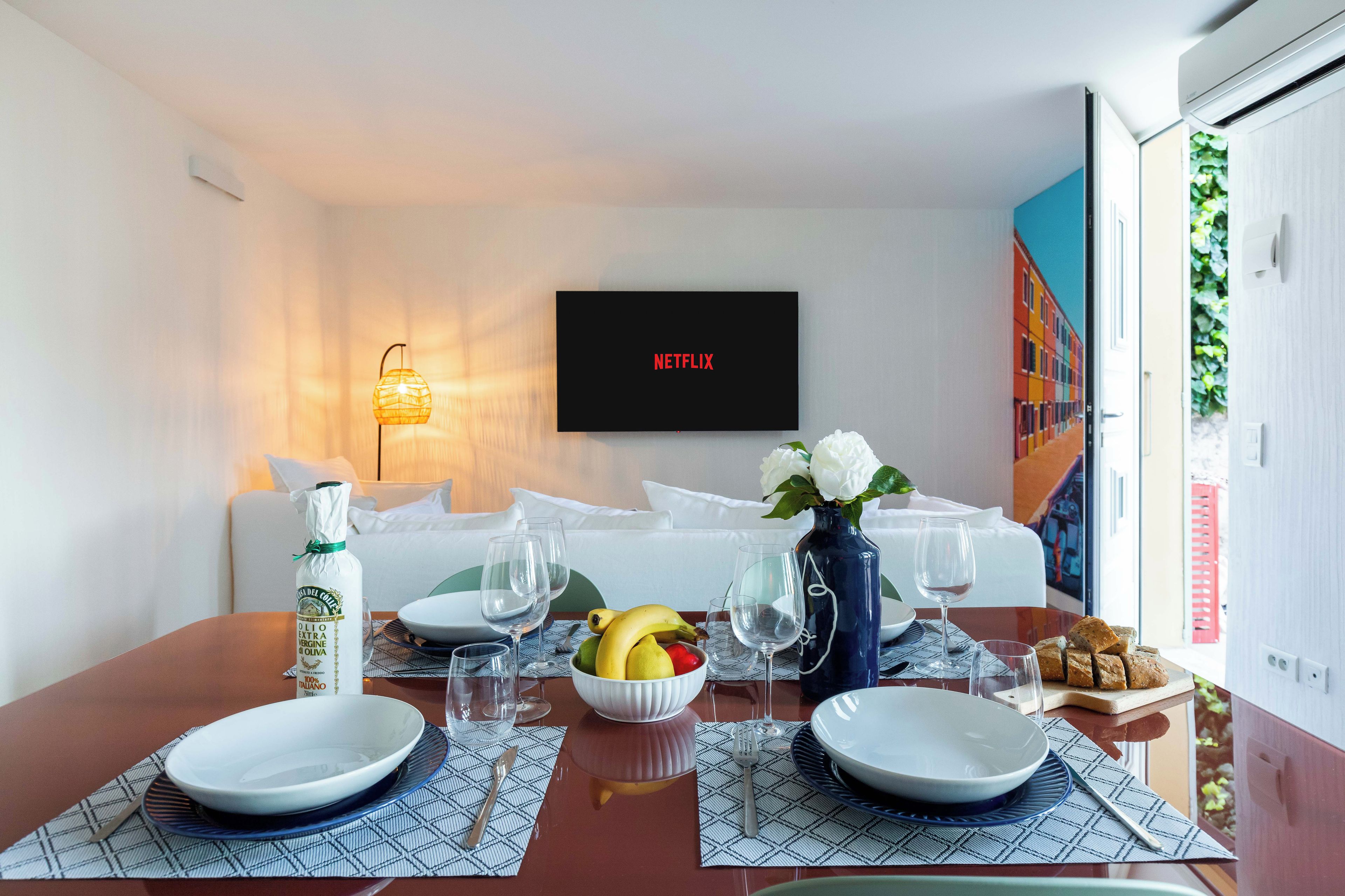 Une chambre d’hôtel avec une table à manger dressée pour deux personnes, une bouteille d’huile d’olive, un saladier de fruits et un vase rempli de fleurs.