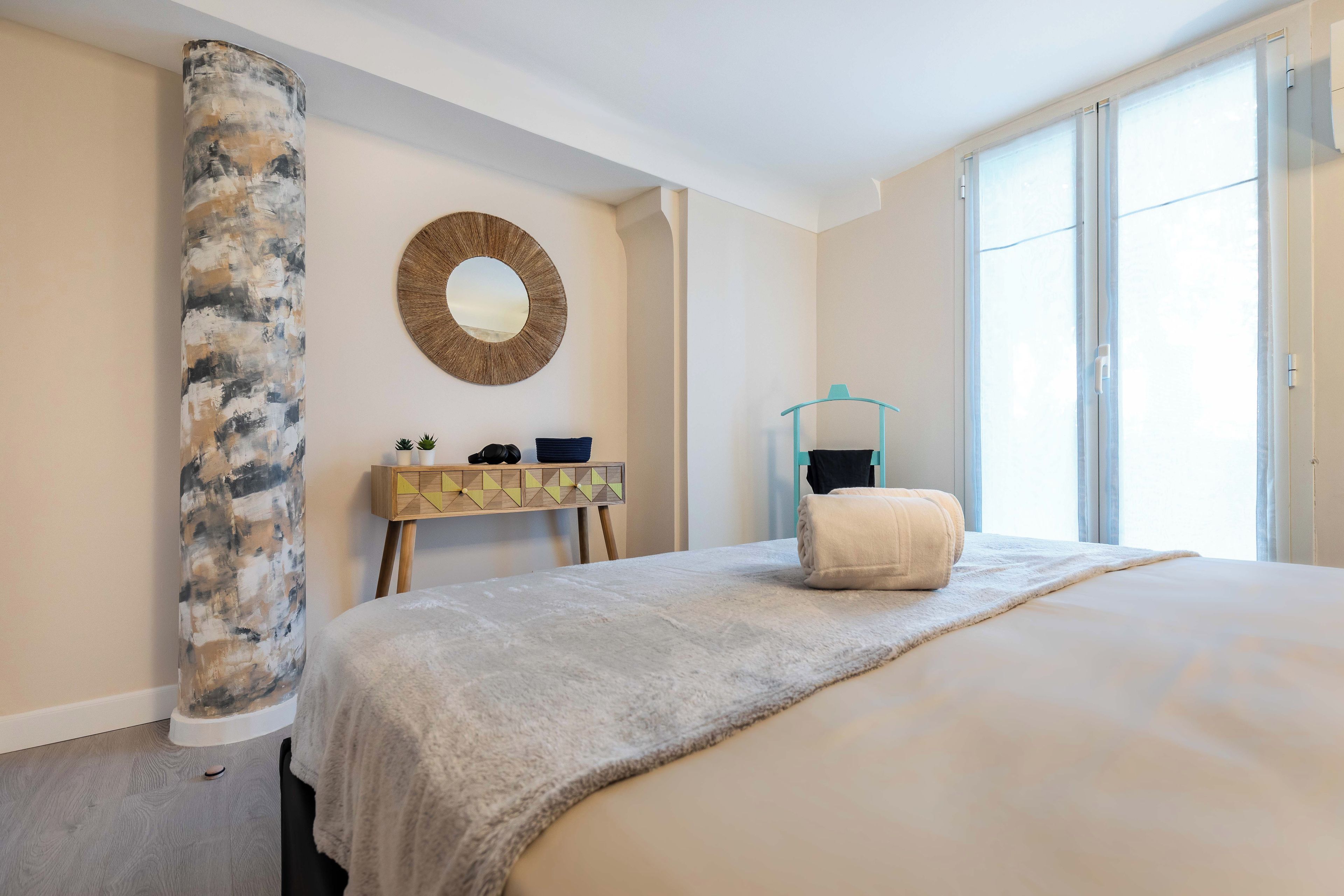 Un dormitorio con una cama grande, un espejo redondo, una mesita de madera y una ventana con cortinas.