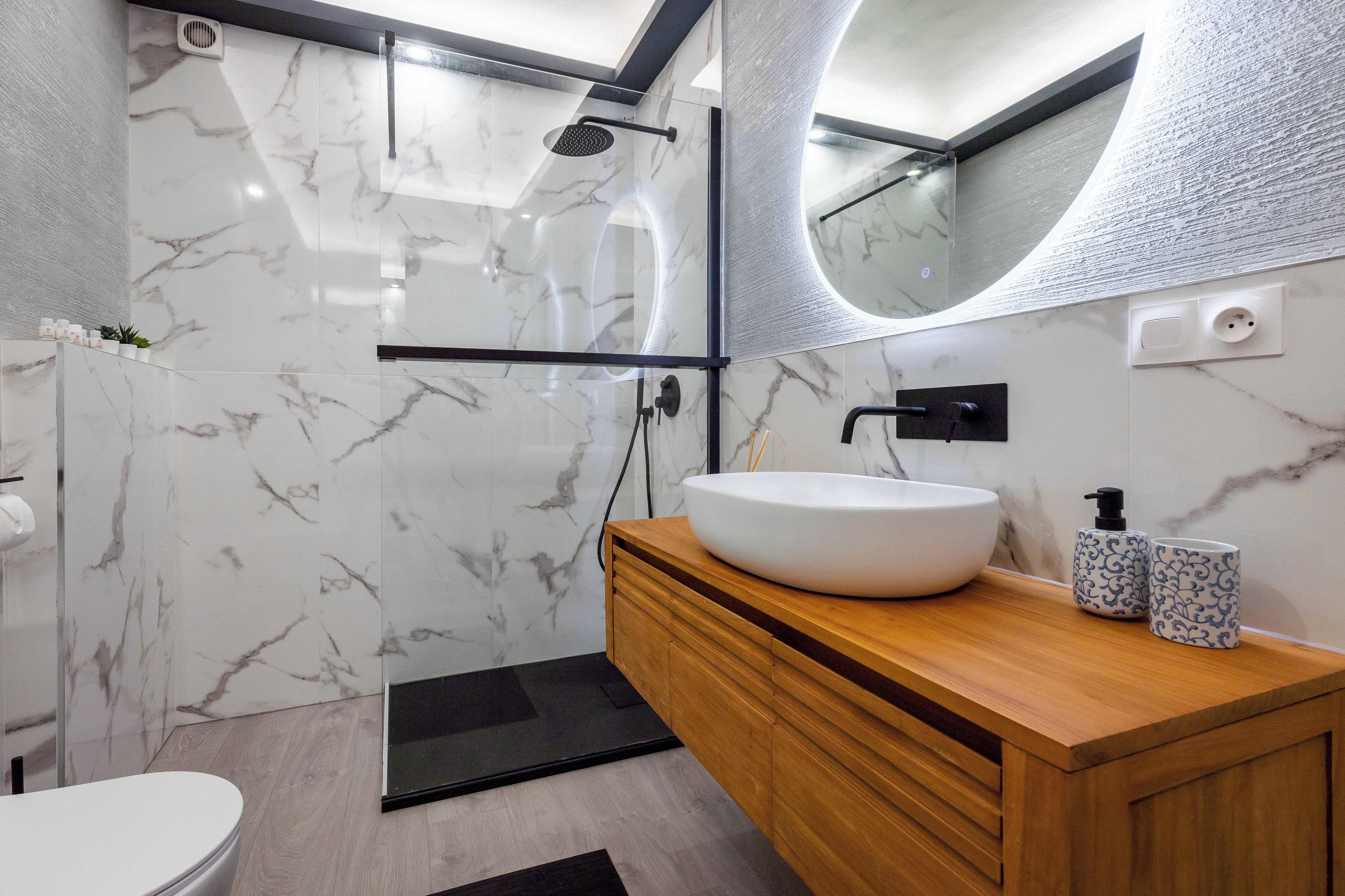 Une salle de bain moderne avec un mur en marbre, un meuble-lavabo en bois, un lavabo blanc et une cabine de douche en verre.