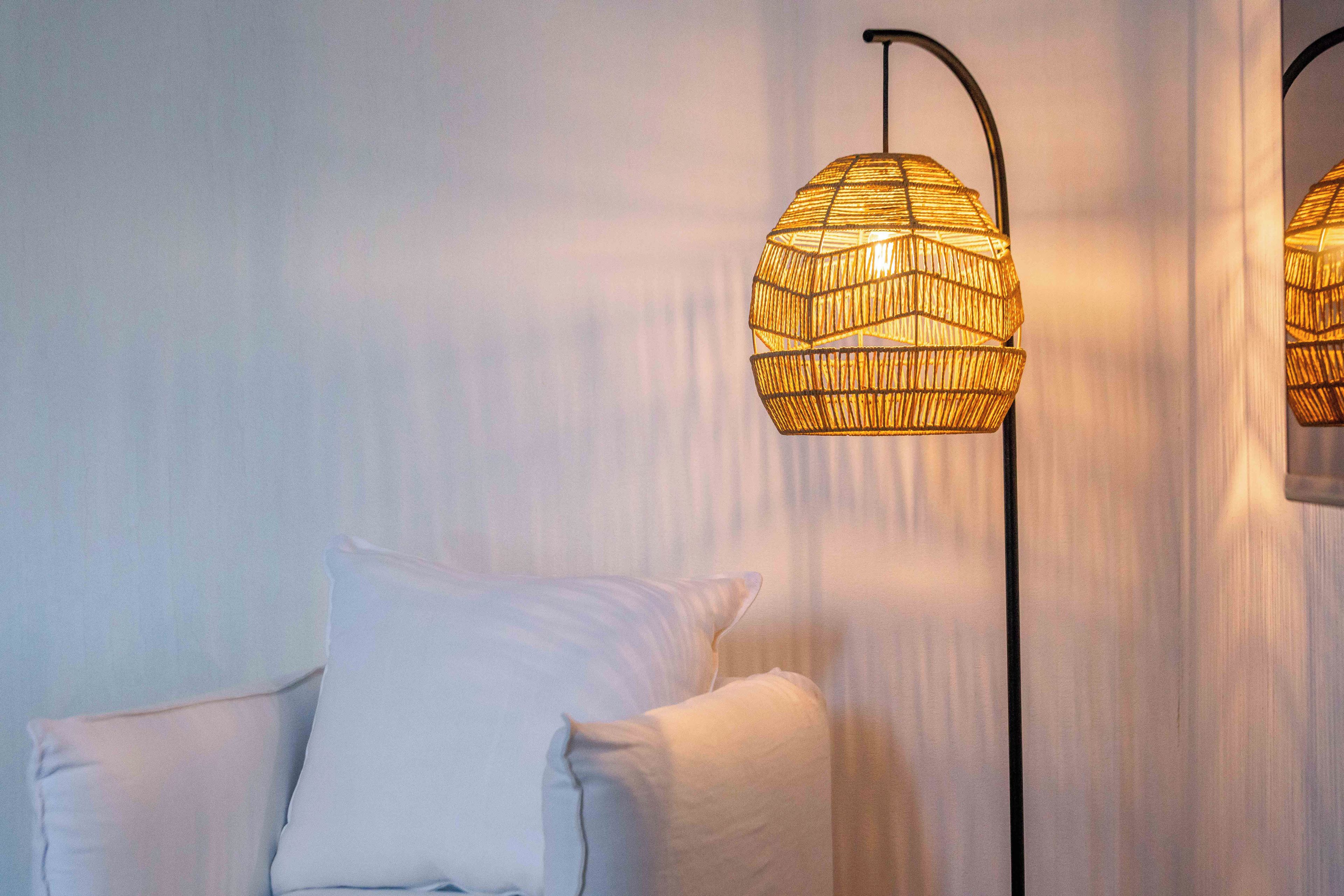 Una habitación con una silla blanca, una lámpara de pie con pantalla tejida y una cortina.