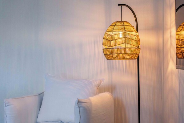 Una habitación con una silla blanca, una lámpara de pie con pantalla tejida y una cortina.