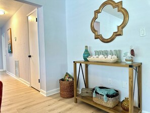 A hallway with a wooden floor, a mirror on the wall, a console table with decorative items, and a wicker basket. - Sarasota 4BR Home Near Siesta Key • Fenced Yard, Fire Pit & Pets (Sarasota)
