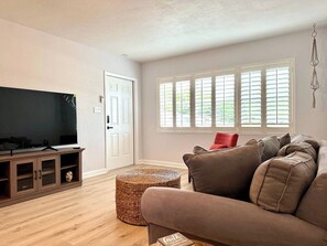 A living room with a flat-screen TV on a wooden cabinet, a brown sofa with cushions, a wicker ottoman, and a window with blinds. - Sarasota 4BR Home Near Siesta Key • Fenced Yard, Fire Pit & Pets (Sarasota)