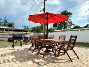 Eine Terrasse im Freien mit einem Holztisch und -stühlen, einem roten Sonnenschirm und einem Grill.