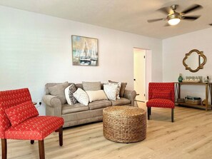 Ein Wohnzimmer mit einer Couch, zwei roten Sesseln, einem runden Korbschuh und einem Deckenventilator, sowie einem Gemälde von Segelbooten.
