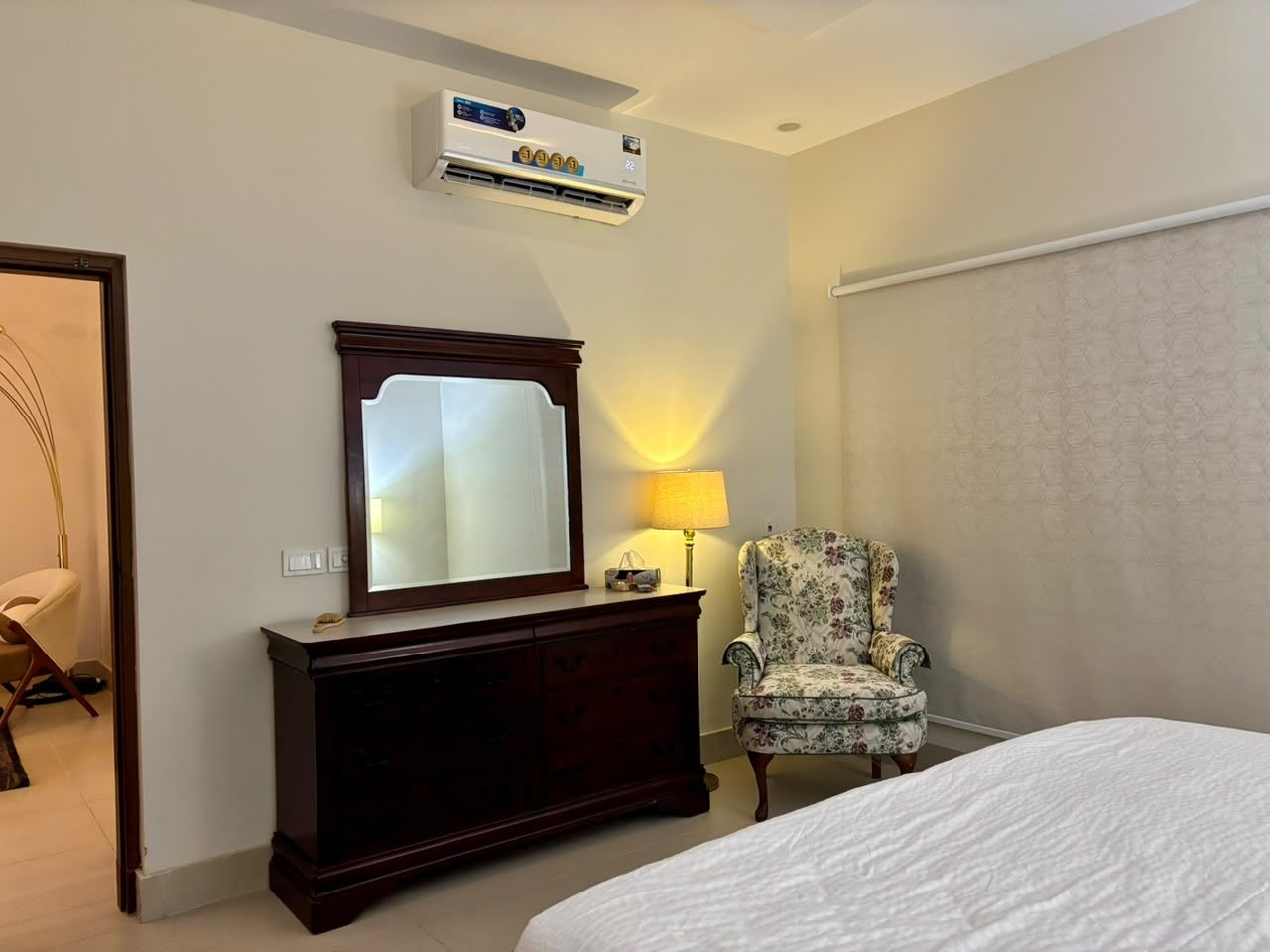 A hotel room with a bed, a dresser, a chair, a mirror, and an air conditioning unit.
