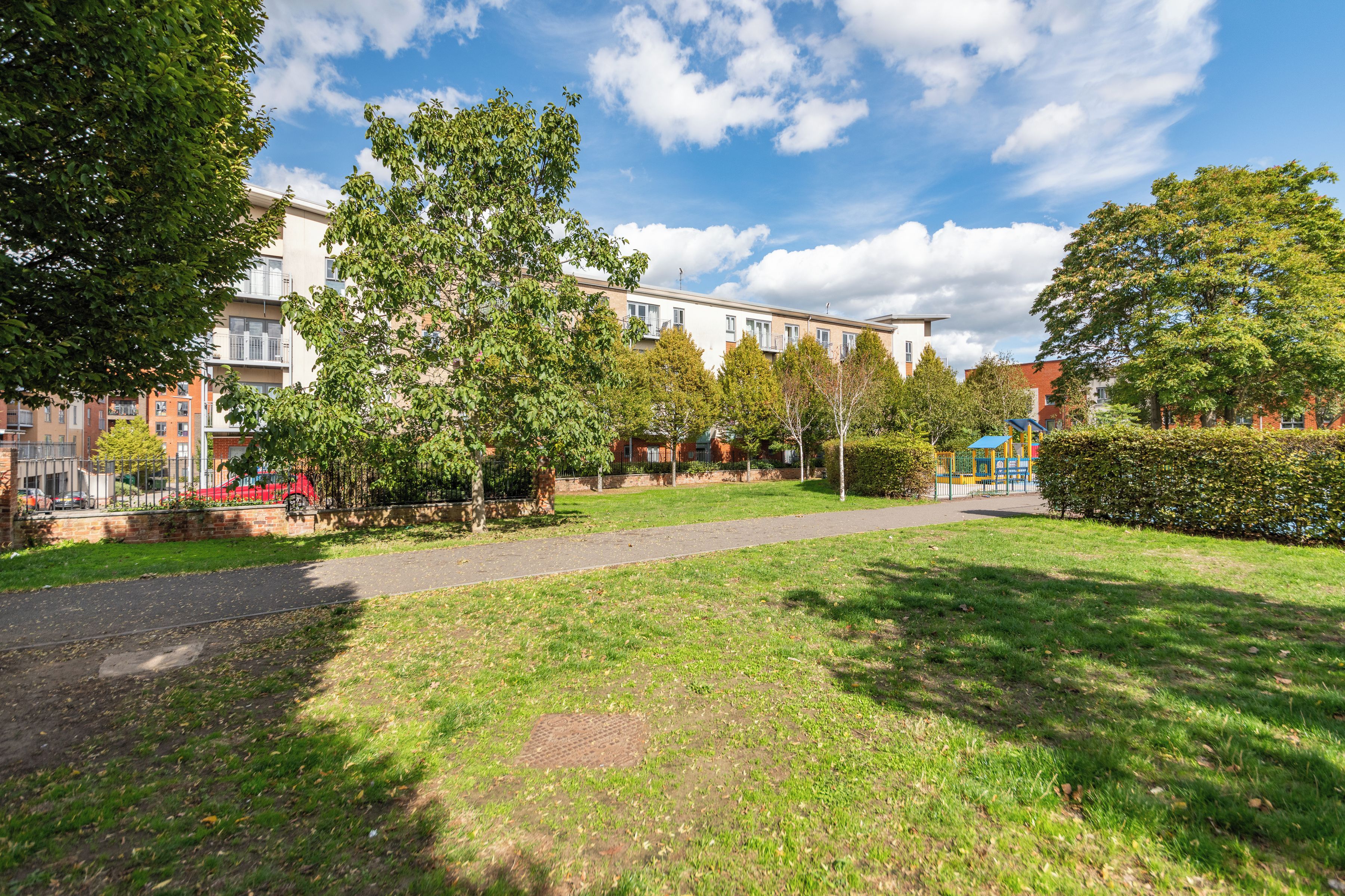 Ein Park mit einem gepflasterten Weg, Bäumen und Wohnhäusern im Hintergrund.