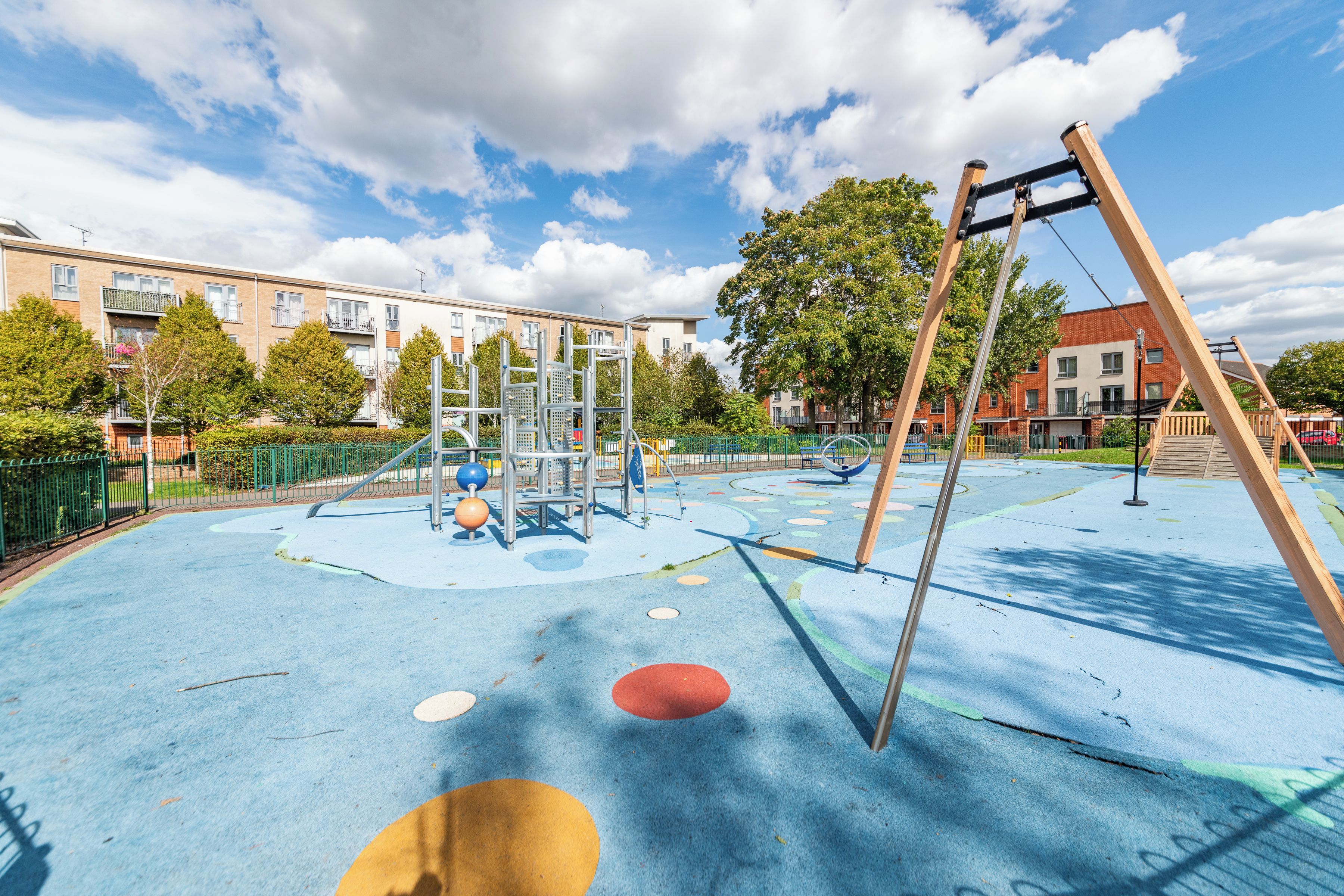 Ein Spielplatz mit Klettergerüst und Schaukeln, umgeben von einem Zaun und Bäumen, mit Gebäuden im Hintergrund.