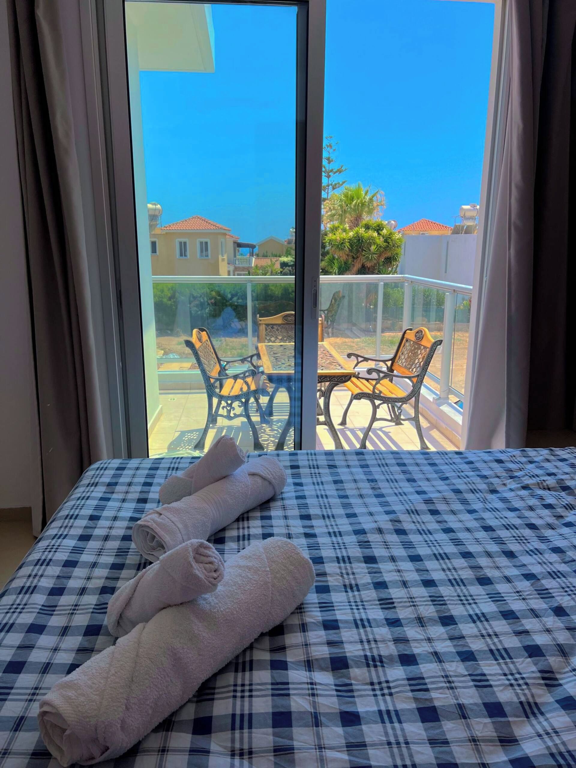 A hotel room with a checkered bedspread, a balcony with a glass railing, and a view of the surrounding buildings and greenery.
