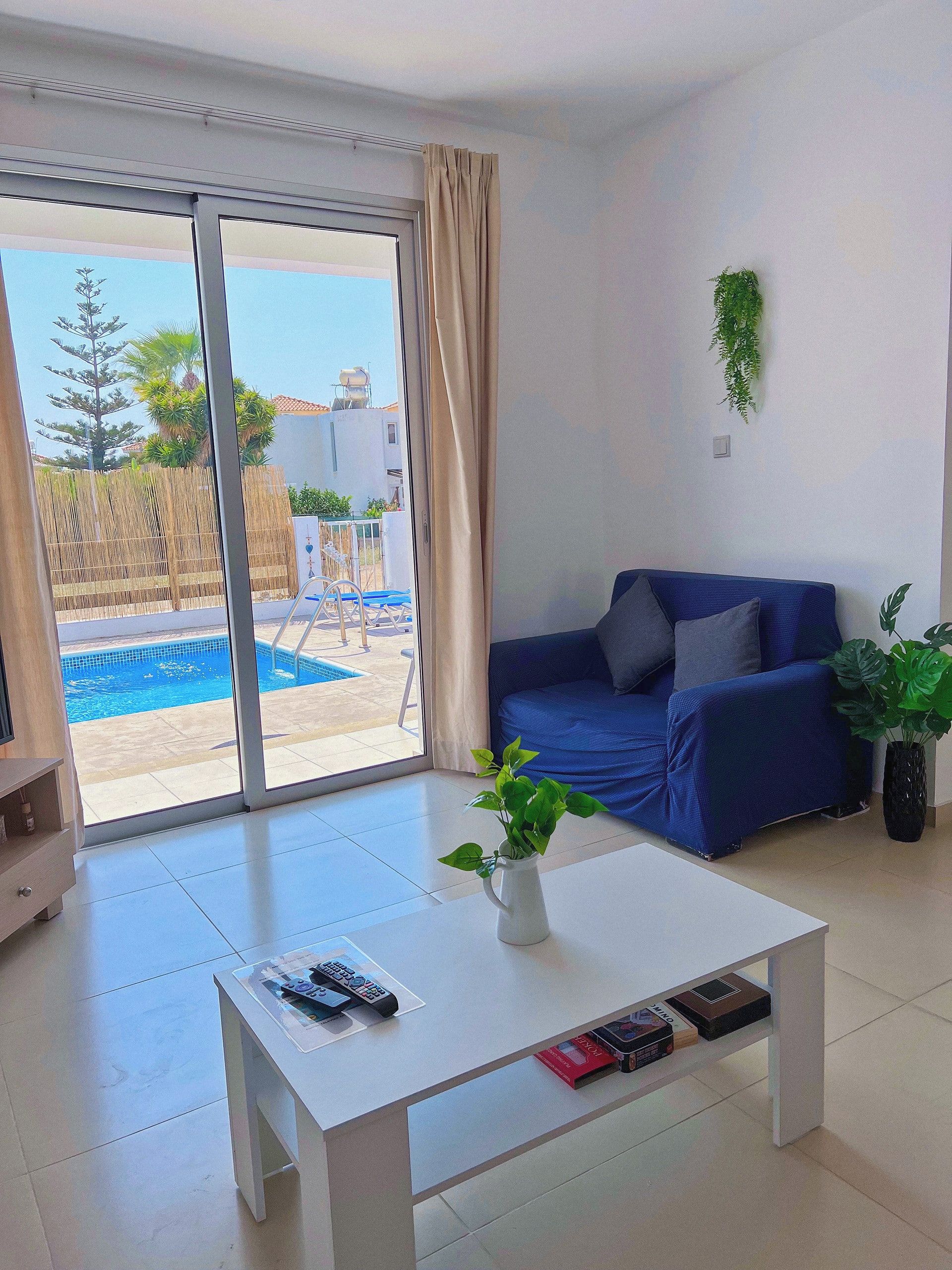 Un salon moderne avec un canapé bleu, une table basse blanche sur laquelle reposent des télécommandes, et une vue sur une piscine à l’extérieur.
