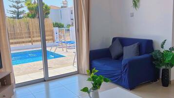 Un salon moderne avec un canapé bleu, une table basse blanche sur laquelle reposent des télécommandes, et une vue sur une piscine à l’extérieur.