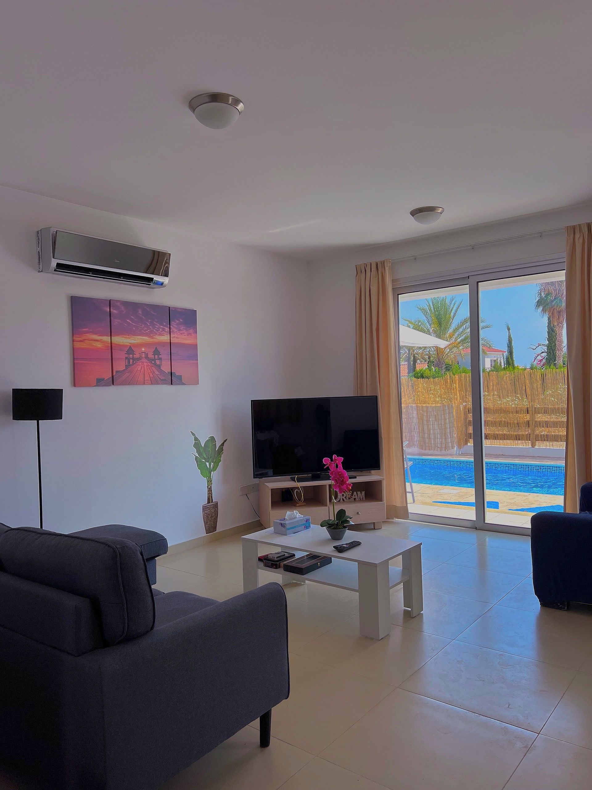 A living room with a flat-screen TV, a sofa, a coffee table, and a view of a swimming pool.