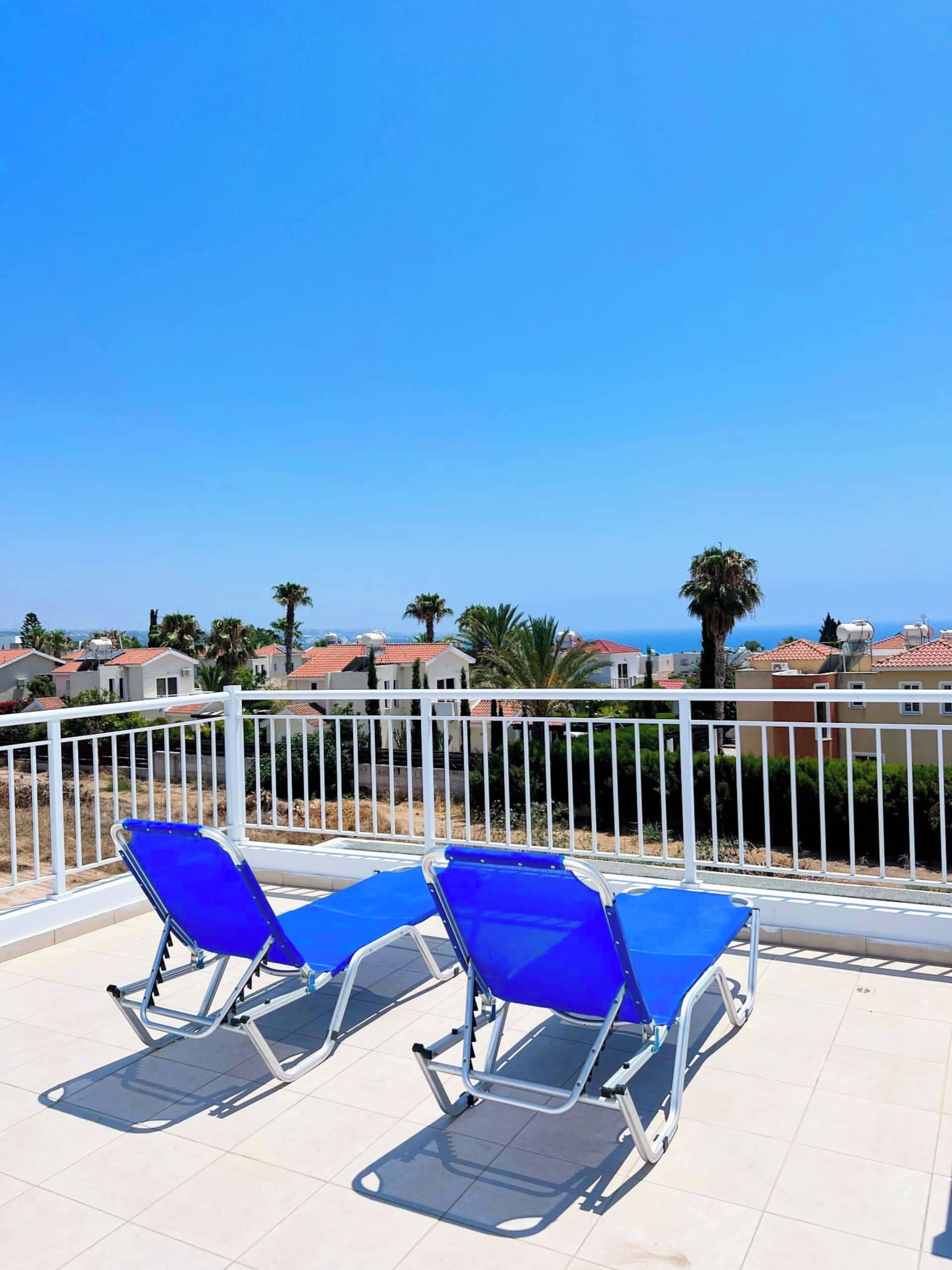A balcony with blue sun loungers and a white railing overlooking a residential area with houses and palm trees.