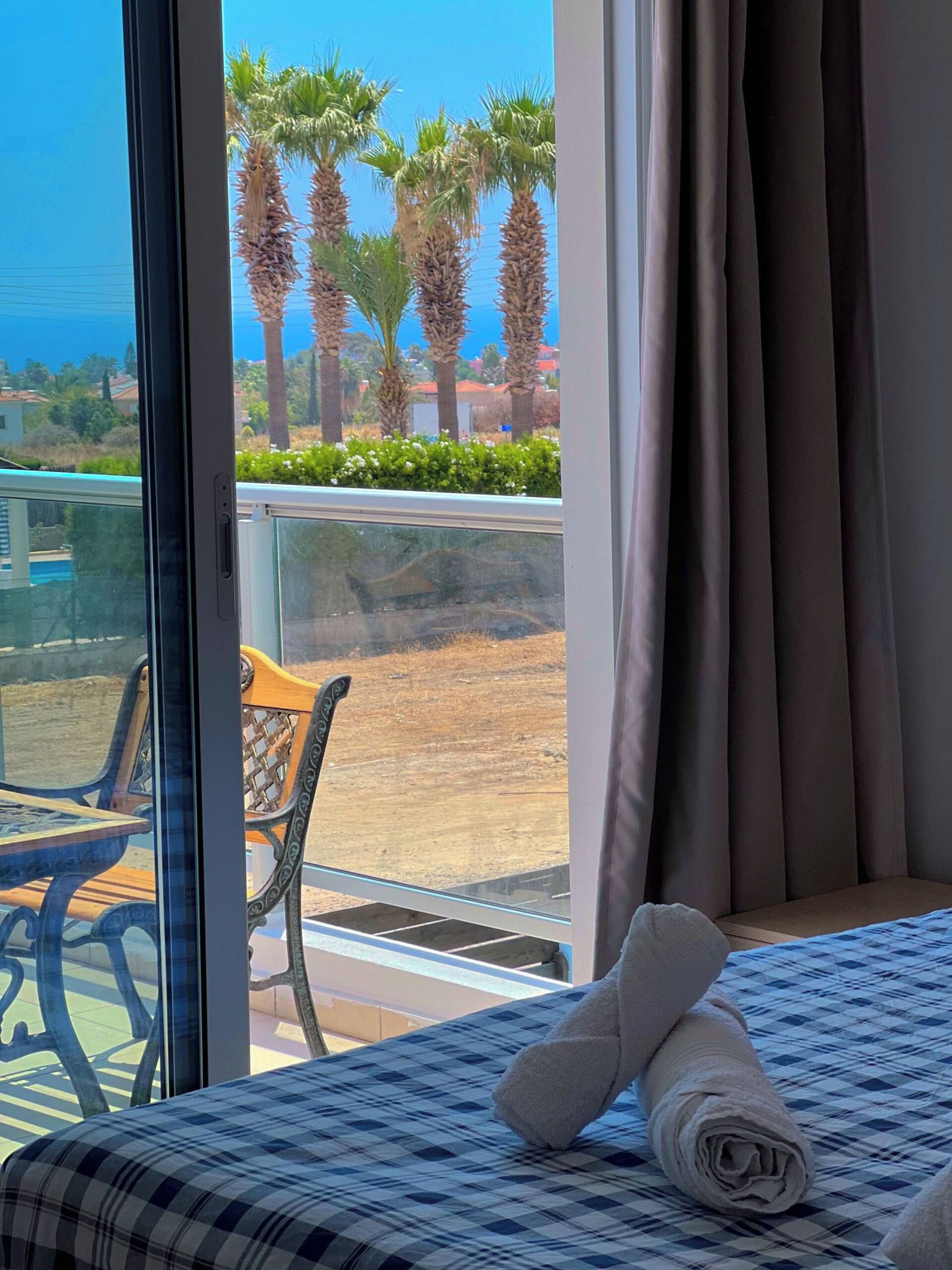 Ein Hotelzimmer mit Bett, Blick auf Palmen und einem Balkon mit Tisch und Stühlen.