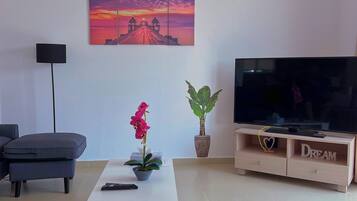 A living room with a flat-screen TV, a white coffee table with a potted plant, a sofa, and a painting of a sunset.
