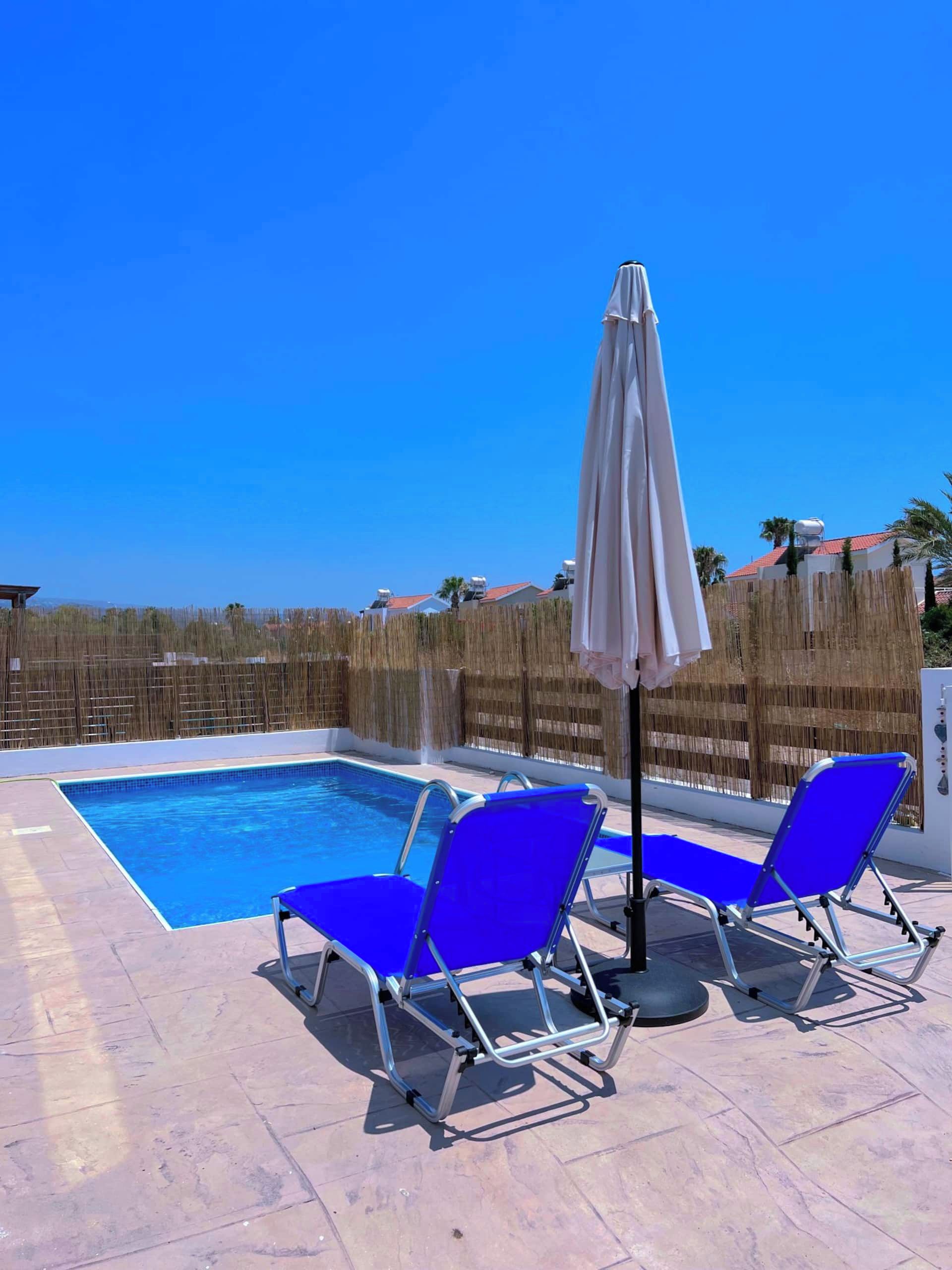 Una piscina con dos sillas reclinables azules y una sombrilla.