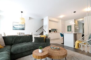 A modern living space with a sofa, two round wooden tables, a hanging chair, and a kitchen area. - Harmony House | Cozy 4-Bedroom Niagara Retreat · EV Charger · Sleeps 8 (Welland)