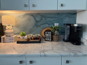 A kitchen counter with a marble backsplash, a coffee maker, a lamp, and decorative items including a sign and plants. - Harmony House | Cozy 4-Bedroom Niagara Retreat · EV Charger · Sleeps 8 (Welland)