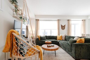 A living room with a green sofa, a macrame chair, a wooden coffee table, and a city view through large windows. - Harmony House | Cozy 4-Bedroom Niagara Retreat · EV Charger · Sleeps 8 (Welland)