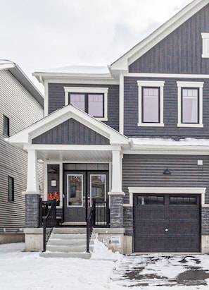 A two-story residential building with a front porch, a garage, and a snow-covered driveway. - Harmony House | Cozy 4-Bedroom Niagara Retreat · EV Charger · Sleeps 8 (Welland)