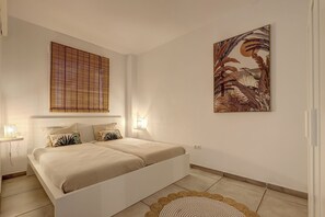 Une chambre à coucher avec un lit, une fenêtre avec des stores, un luminaire fixé au mur et un tableau encadré.