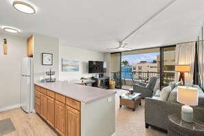 A modern apartment with a kitchen, dining area, and living room. It features a large window with a view of the ocean and a balcony with outdoor seating. - Sugar Beach PH08 | Penthouse Ocean Views, Beachfront Pool & Central A/C (Kihei)