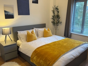 A neatly made bed with white linens and a yellow throw, a bedside lamp, a small bedside table, and a potted plant. - Luxury 2-Bed 2-Baths City Centre Apartment |  FREE Secure Parking, Balcony (Birmingham)