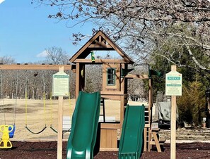A playground with slides and swings, surrounded by trees and a wooden playhouse.