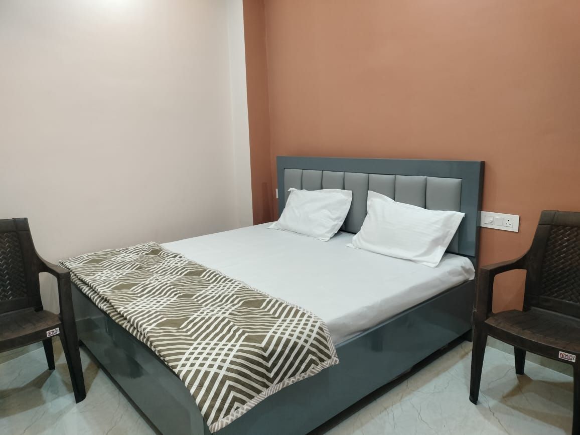A single bed with a patterned bedspread and two pillows, flanked by two chairs, in a room with a plain wall and a visible electrical outlet.