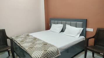 A single bed with a patterned bedspread and two pillows, flanked by two chairs, in a room with a plain wall and a visible electrical outlet.