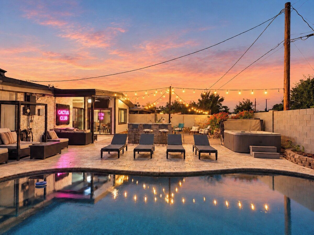 A pool area with lounge chairs and a pool, surrounded by a building with a neon sign, under a sunset sky.