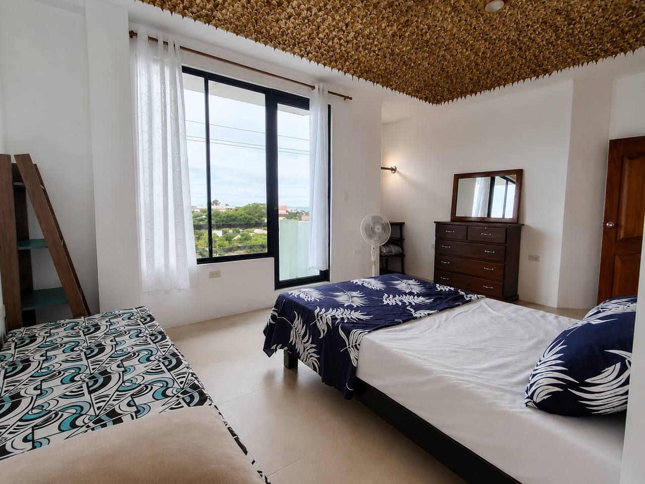 A bedroom with two beds, a window with curtains, a fan, a dresser, and a wooden ladder.