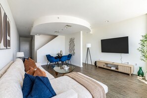 Een moderne woonkamer met een gebogen plafond, een flatscreen televisie aan de muur, een eethoek met blauwe stoelen en een bank met kussens.