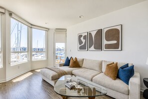 A modern living room with a large window, a glass coffee table, and a beige sofa with cushions. - Coral Marina' Water Views Pool Jacuzzi Sauna Gym (Marina del Rey)
