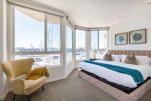 Ein Hotelzimmer mit großem Fenster und Blick auf eine Marina, einem Bett mit Kopfteil, einem Sessel mit Überwurfdecke sowie zwei gerahmten Kunstwerken an der Wand.