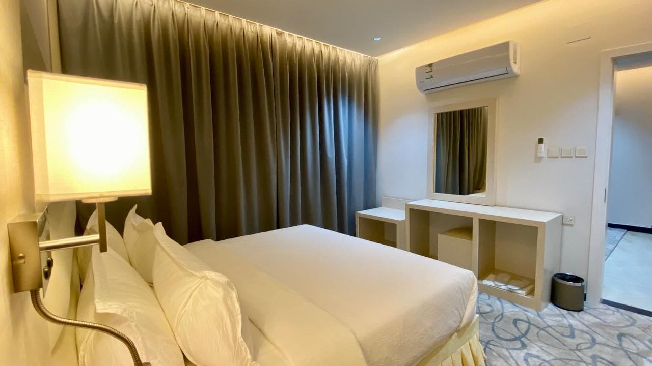 A hotel room with a bed, a desk, a mirror, an air conditioning unit, and a window with curtains.