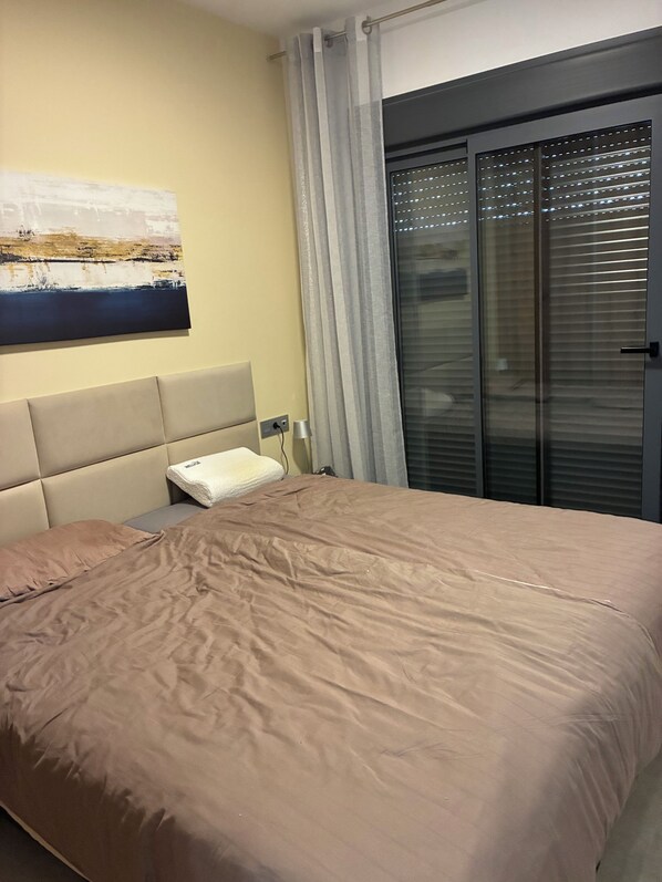 A bedroom with a bed, a headboard, a bedside table, a lamp, and a window with blinds.
