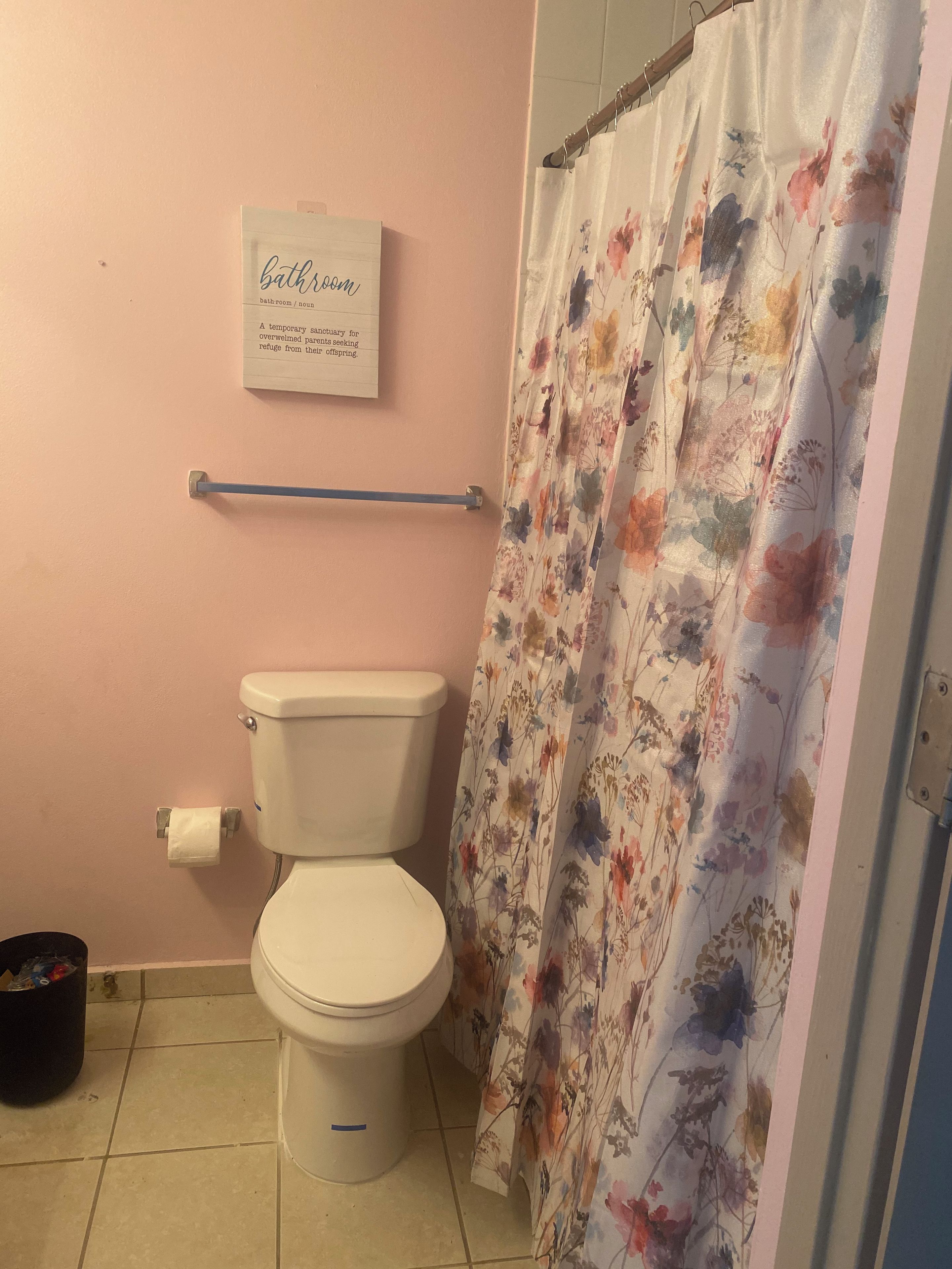 Casa de banho com sanita, rolo de papel higiénico, cortina de chuveiro com padrão floral e um aviso na parede.