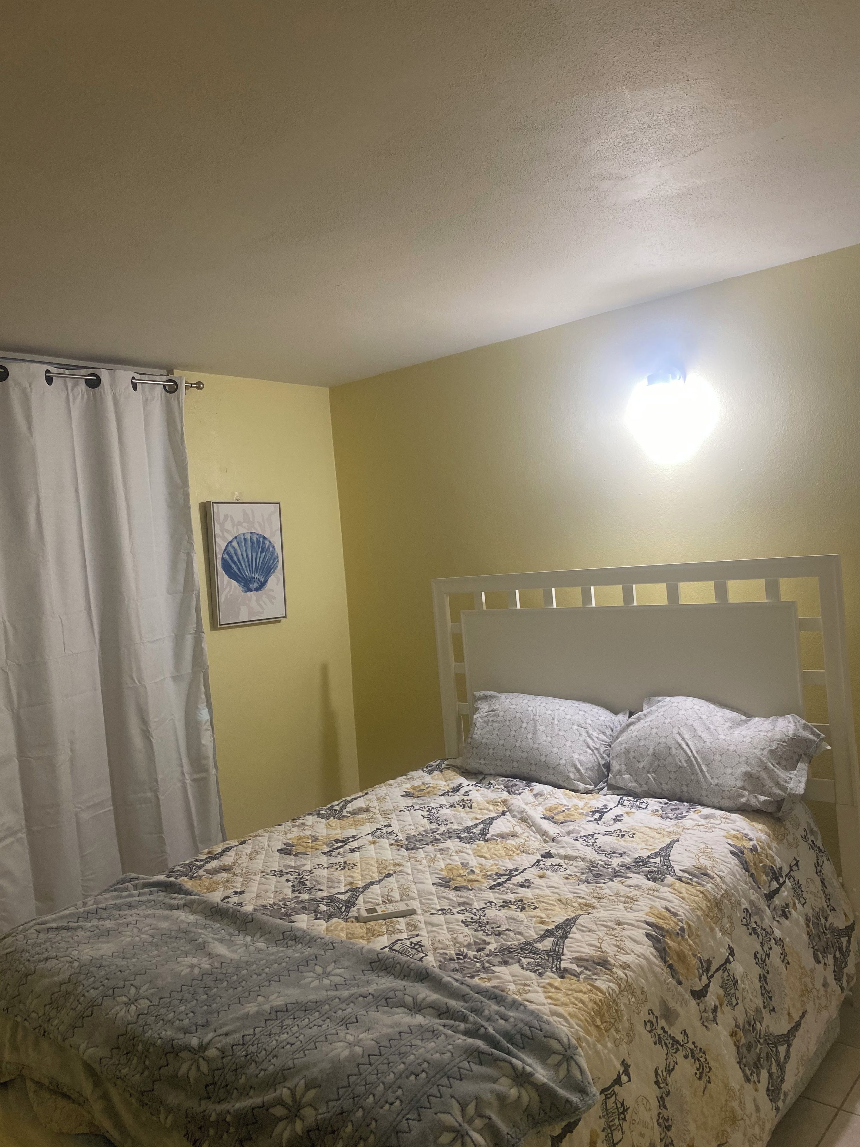 Quarto com cama, cabeceira branca, luminária na parede e cortina.