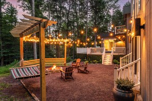 An outdoor patio area with a wooden pergola, string lights, a fire pit, and Adirondack chairs. - 6BR Poconos Home - Indoor Heated Pool & Hot Tub - Theater, Gym and Game Room (Cresco)