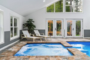 A pool area with lounge chairs, a hot tub, and a view of the outdoors through large windows. - 6BR Poconos Home - Indoor Heated Pool & Hot Tub - Theater, Gym and Game Room (Cresco)