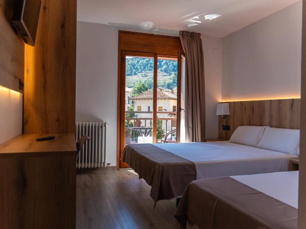 Een hotelkamer met twee bedden, een houten hoofdbord, een radiator en uitzicht op een dorp door een groot raam.