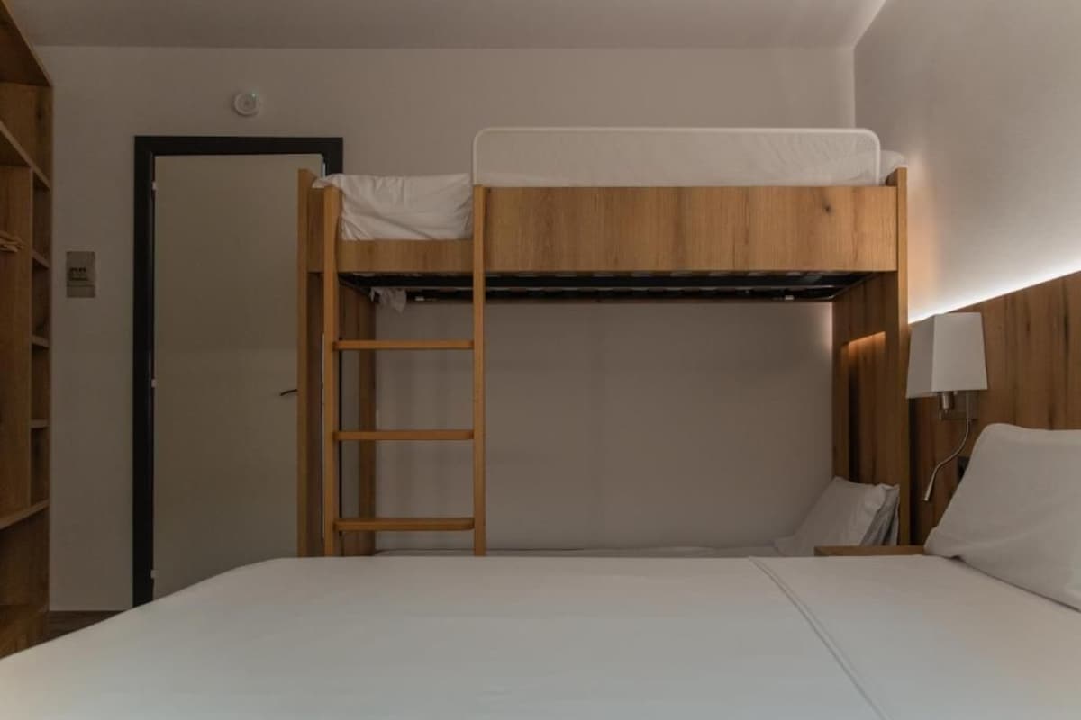 Una cama litera con escalera de madera, un colchón blanco y una mesita de noche con una lámpara.