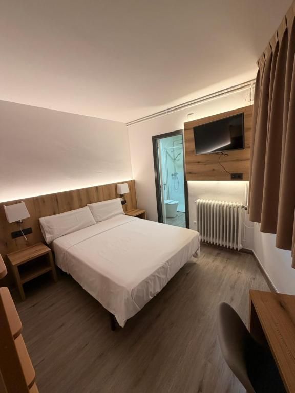 A hotel room with a bed, bedside tables, a desk, a chair, a TV mounted on the wall, and a window with curtains.